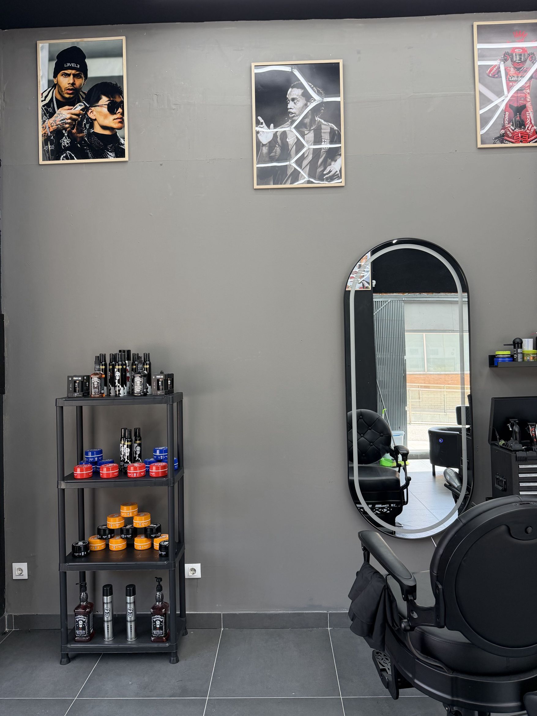 Pared gris con tres carteles enmarcados, una estantería negra con pomadas y botes de peinado, y un espejo de arco junto a una silla de barbero.