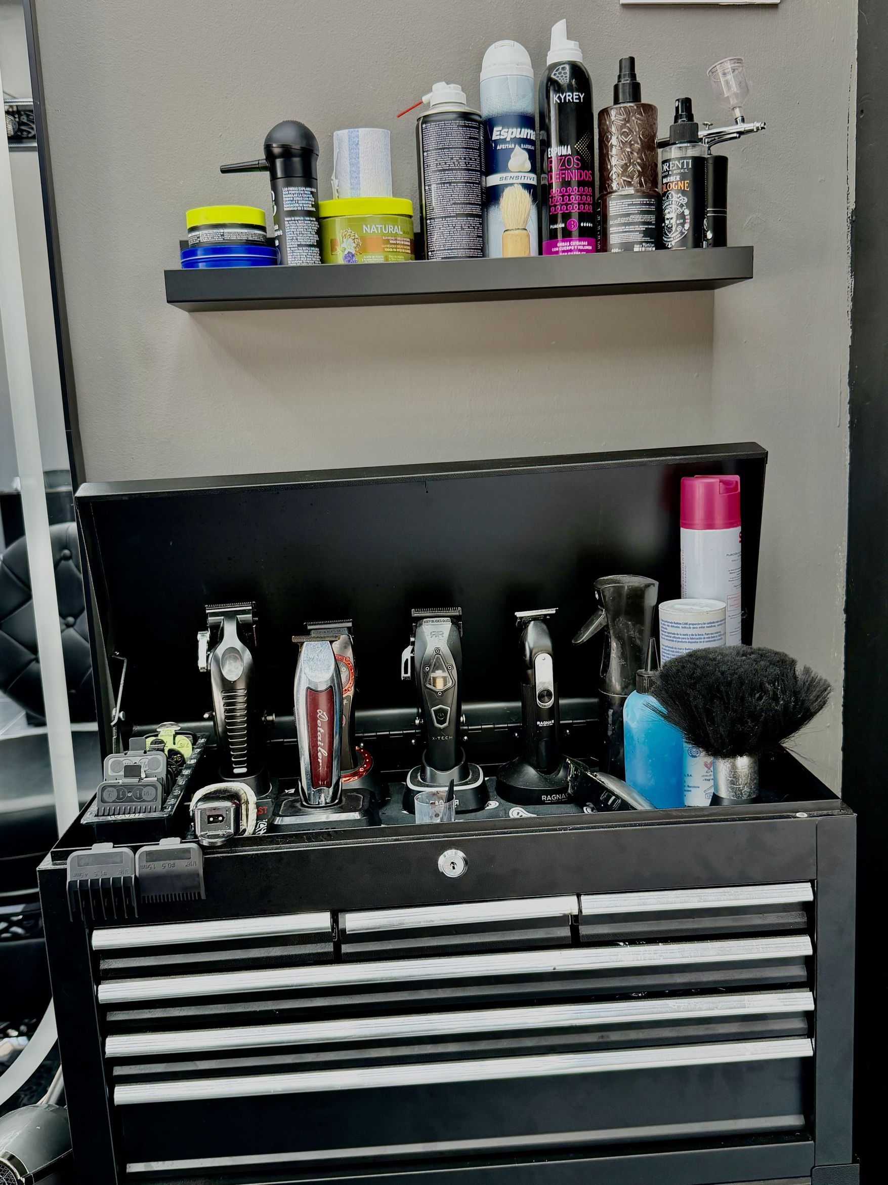 Estante con sprays y productos de peinado sobre un maletín negro abierto con cinco máquinas de cortar, una brocha de afeitar y un bote de polvos.