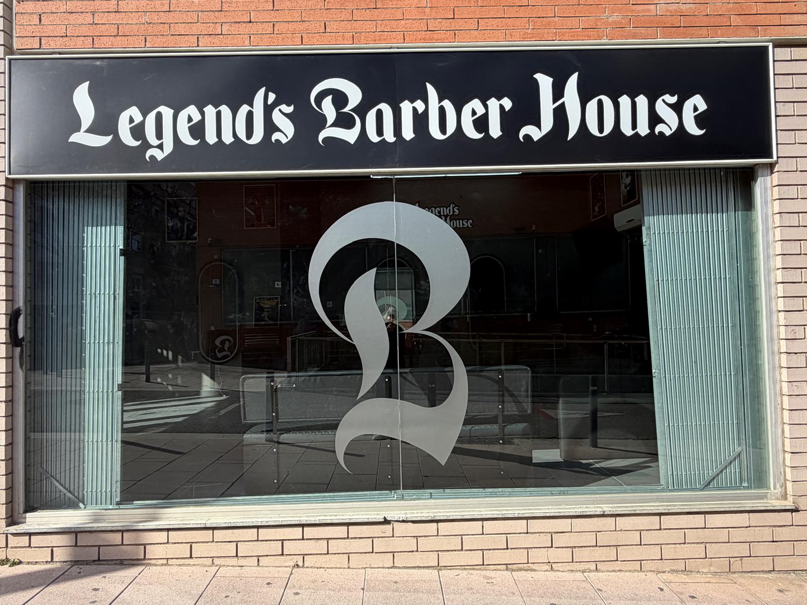 Fachada de la barbería: rótulo negro con el nombre Legend's Barber House en letra gótica sobre el escaparate, monograma B en el cristal y ladrillo rojo y beige alrededor.