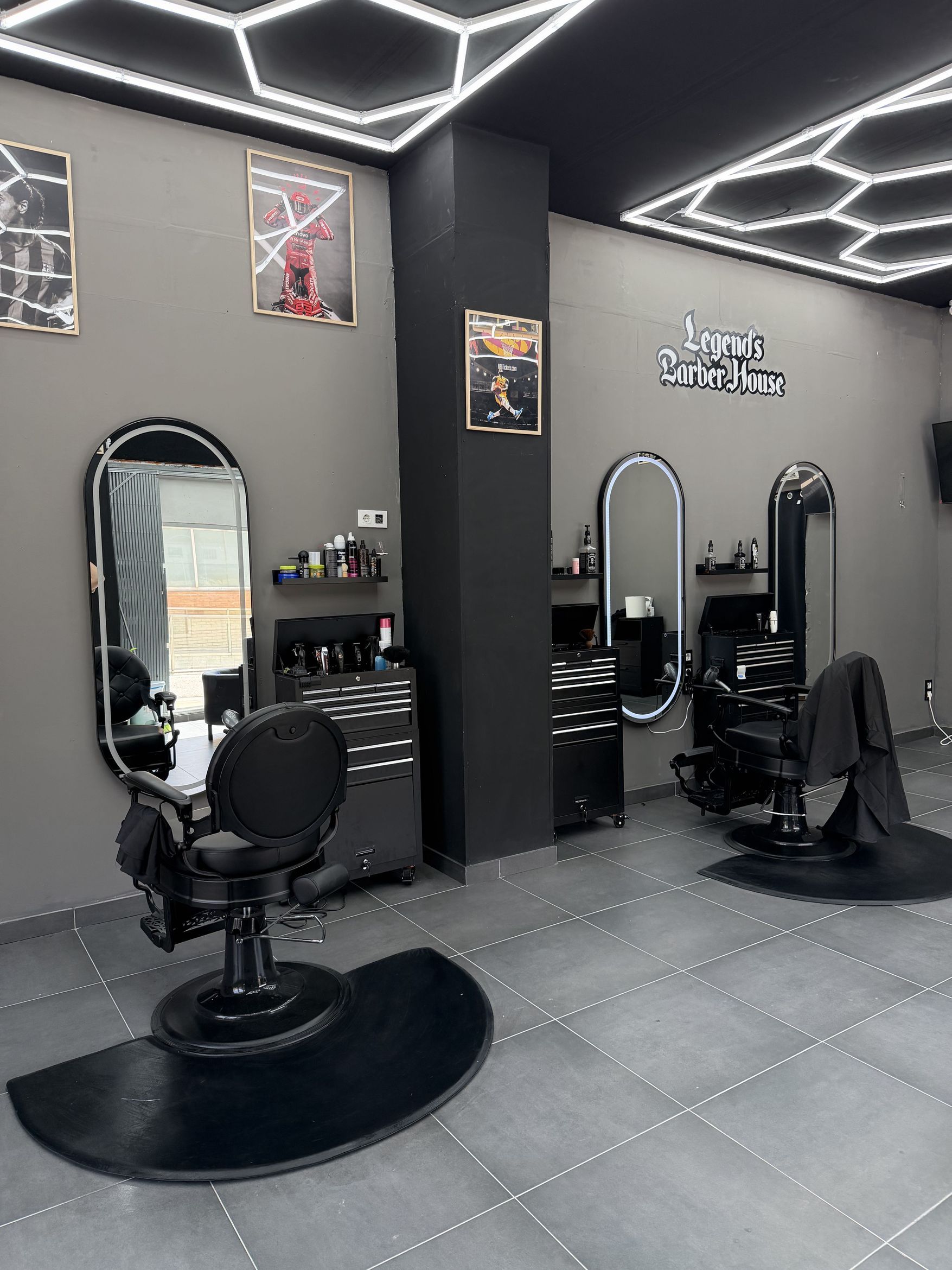 Interior de la barbería con dos puestos de trabajo, sillas negras sobre alfombrillas, espejos de arco iluminados, techo de paneles LED hexagonales y el logotipo de Legend's Barber House pintado en la pared gris.