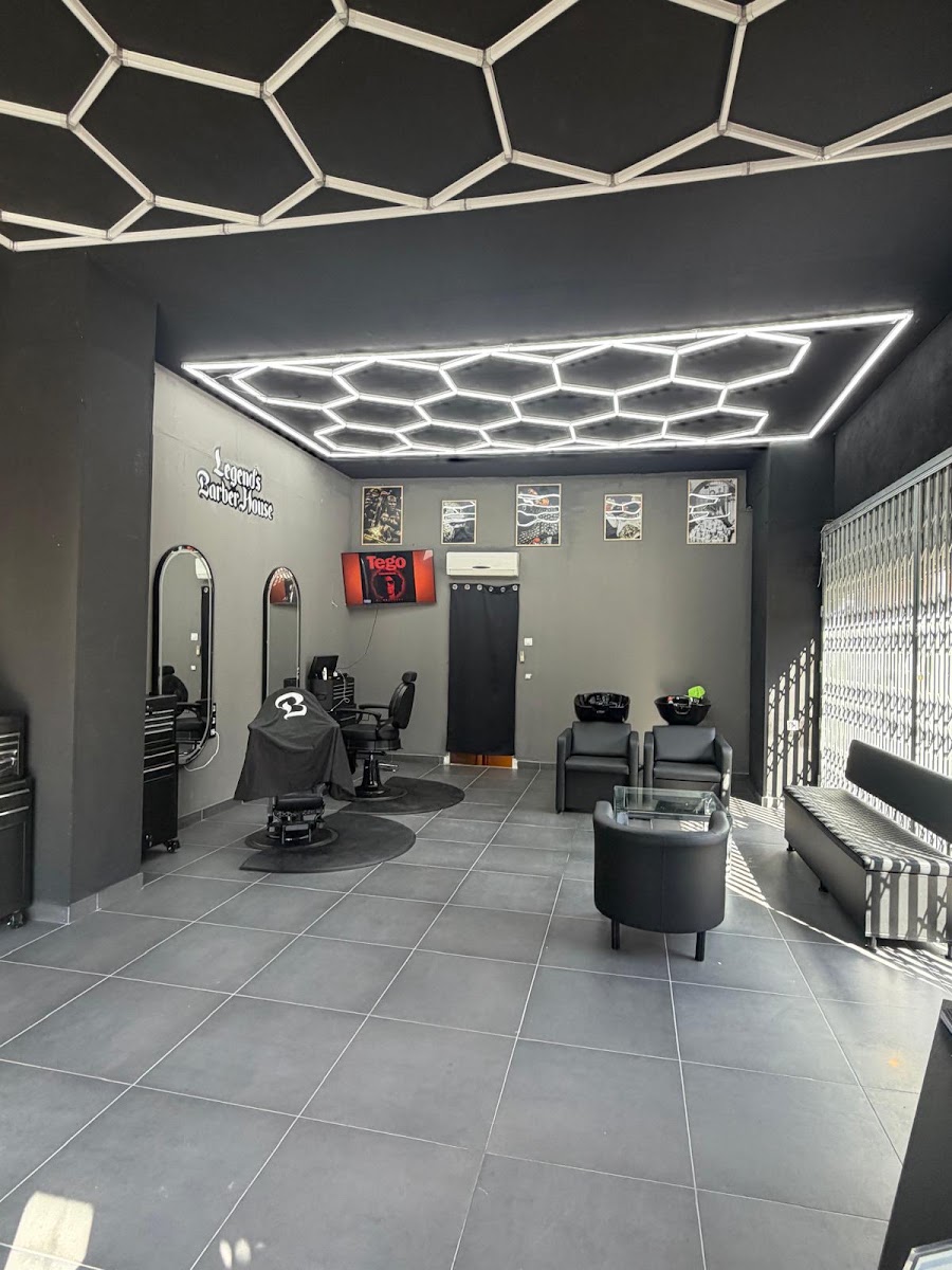 Interior de la barbería hacia el fondo: techo de paneles LED hexagonales, suelo de baldosa gris, una silla de barbero cubierta con una capa negra, dos lavacabezas y un banco de espera.