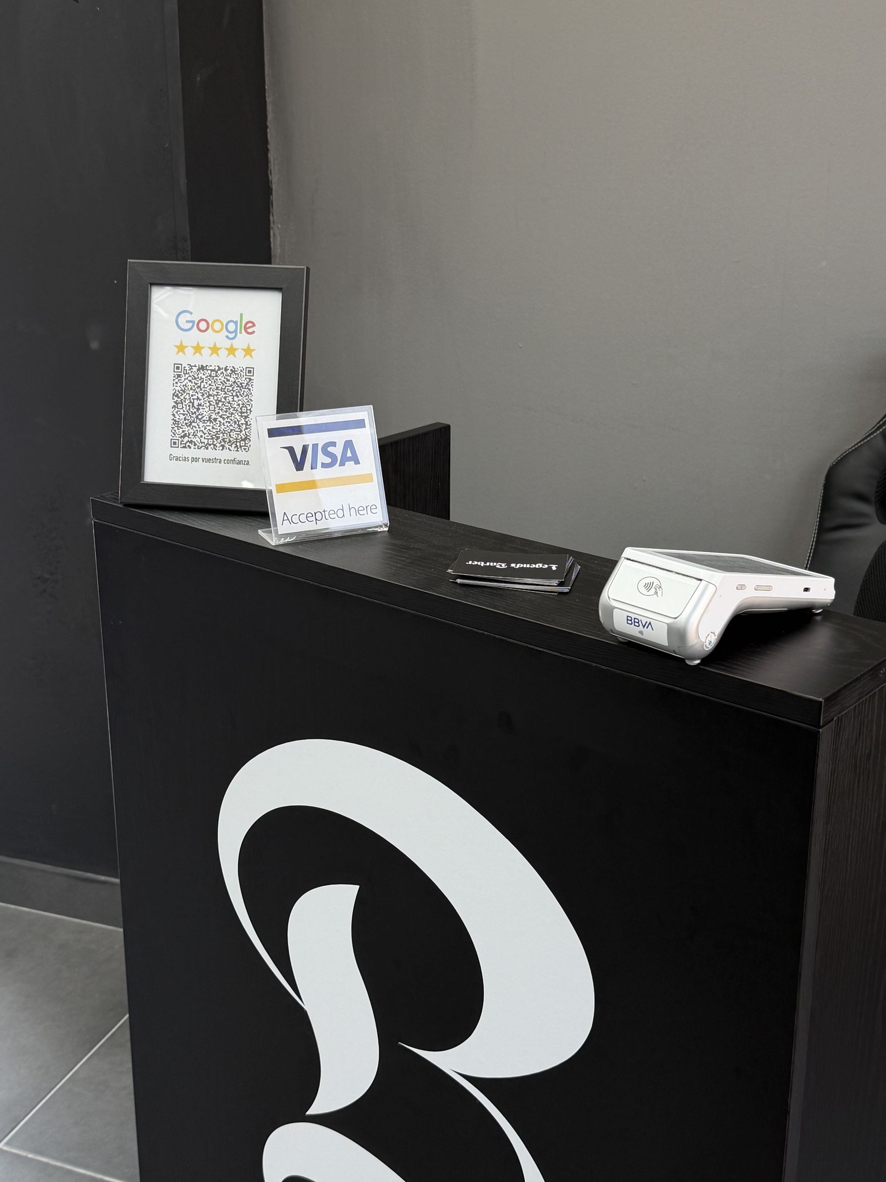 Mostrador de recepción de madera negra con el monograma B en blanco, un cartel de VISA aceptada, tarjetas de visita y un datáfono.